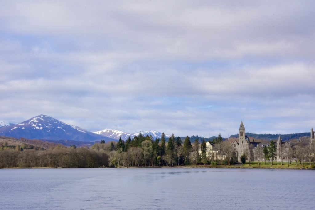 across a wide loch, greenery with a church spire poking out above the trees and a snowy mountain in the background of this classic Scottish scene