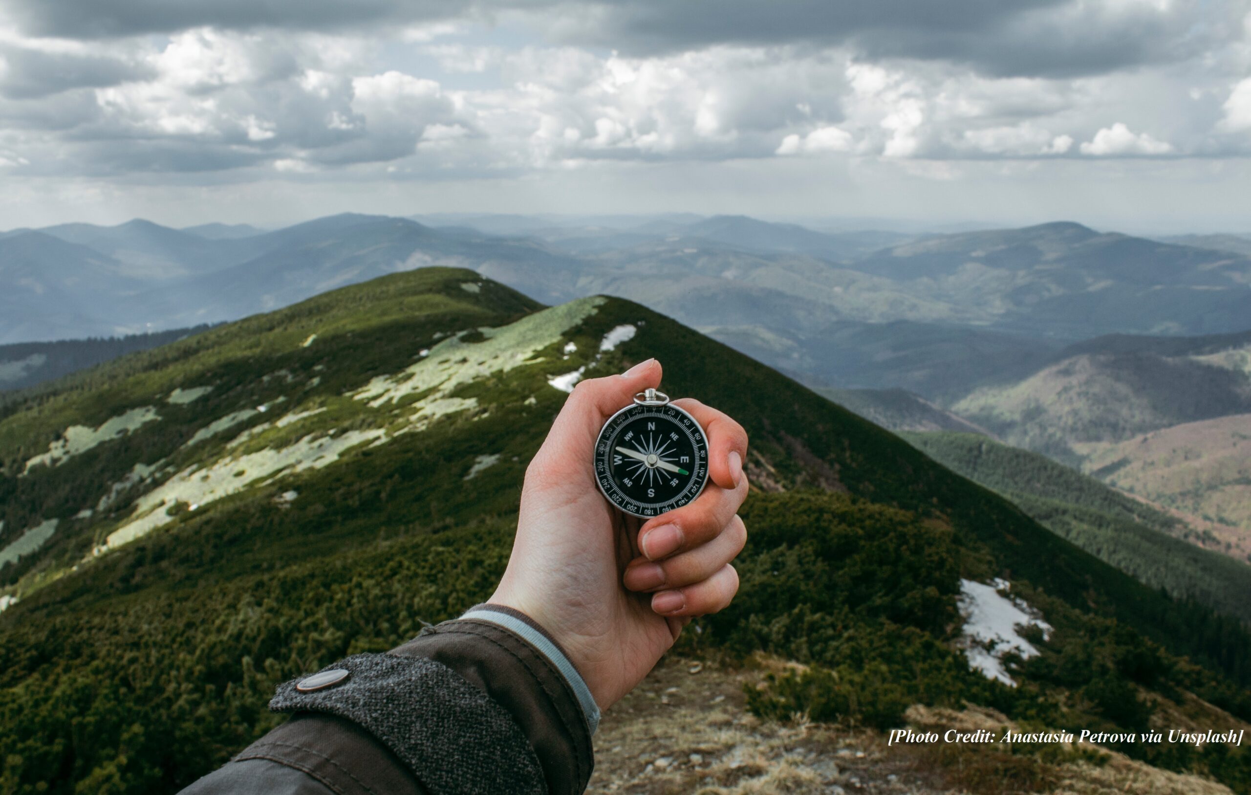hand holding a compass in front of a green mountain-range