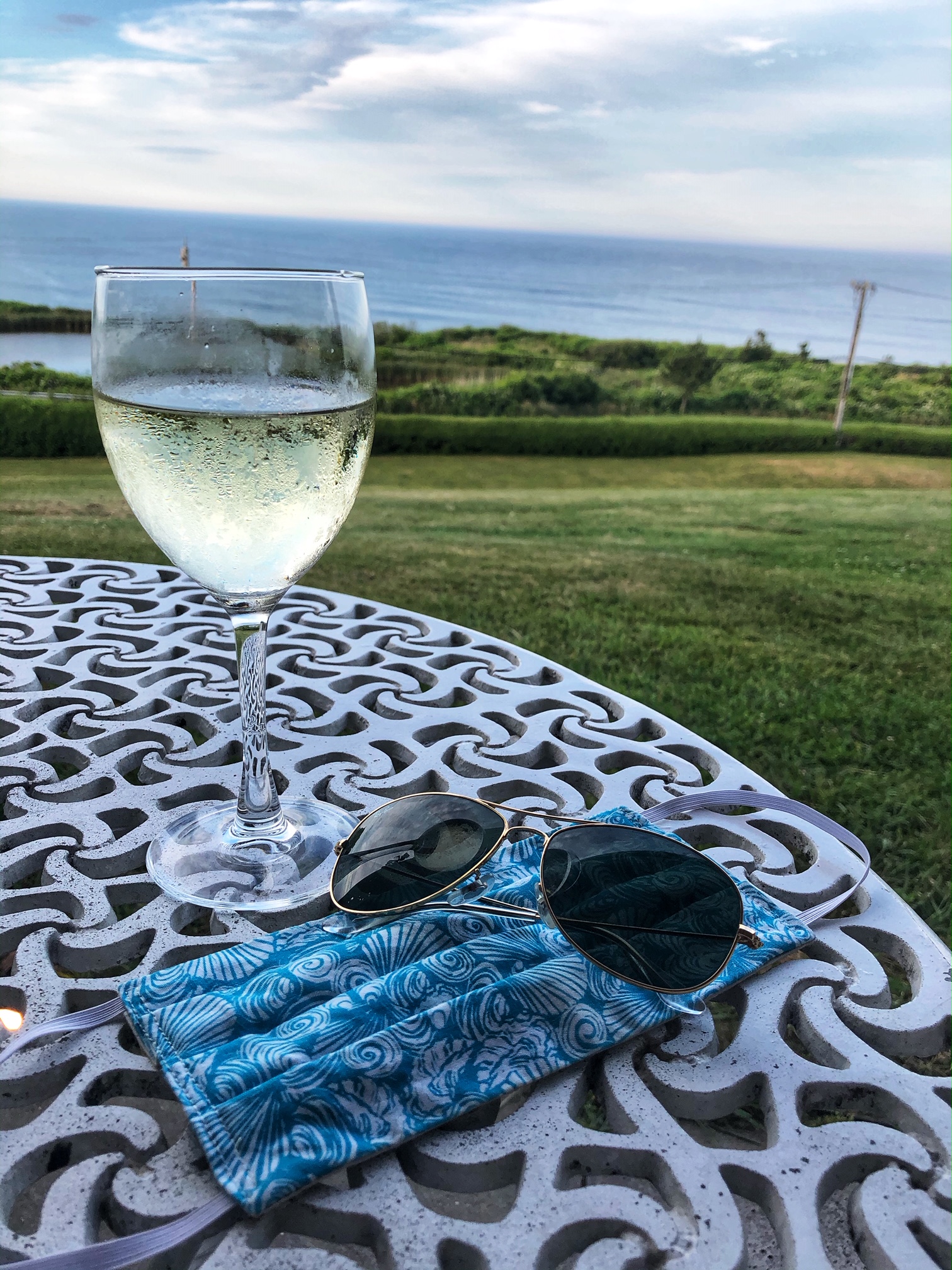 a glass of wine, a homemade mask and sunglasses on a white table with a bay in the background in Rhode Island