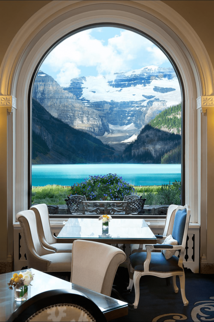 a dining room table with a view of the stunning blue waters and huge snow-capped mountains at the Fairmont Chateau Lake Louise in the Canadian Rockies