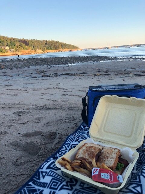 to-go food on a blue blanket with the sand, sea and pine trees in the background a picnic on Seal Beach in Maine
