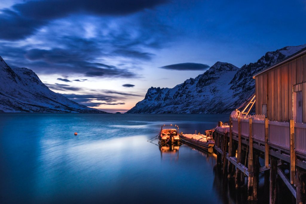 the side of a cabin, a small boat docked with lights and sunrise over the bay and snow mountains in Norway