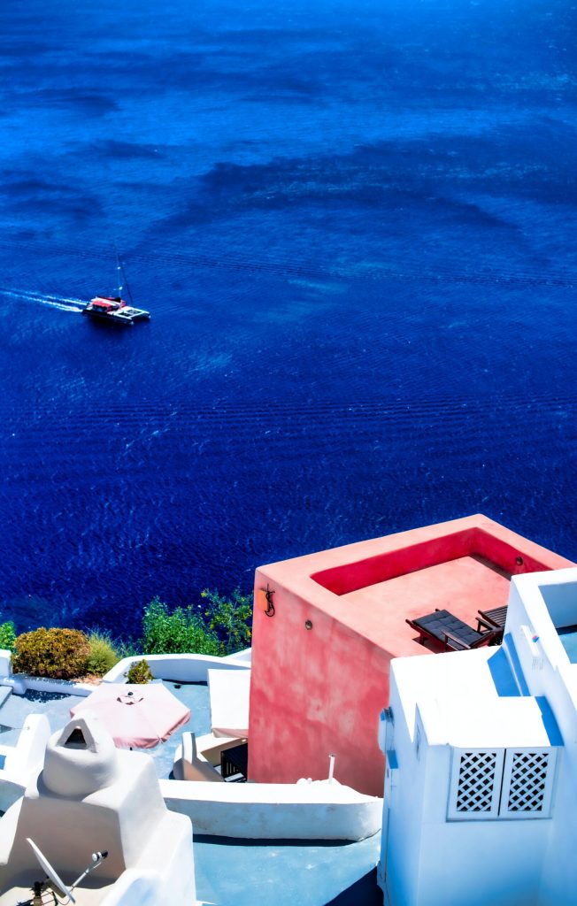 white and pink buildings overlooking a yacht sailing in cobolt blue water in Greece