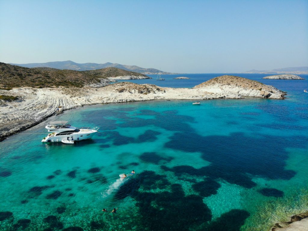 aerial view of people swimming next a yacht in turquoise blue waters and white rocks in the Greek Islands