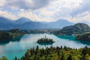 view of Lake Bled from atop a hill looking down at the blue lake with small island and surrounded by green looming mountains in Slovenia