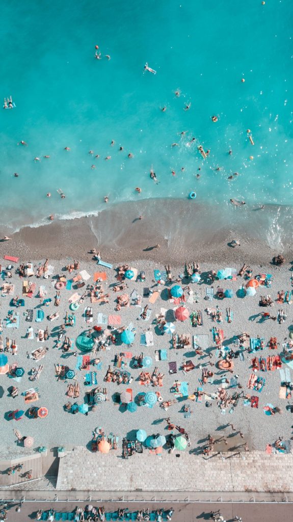 aerial view of a beach with the light blue water on top and the grey pebbles covered with people on towels and pastel colored umbrellas in Page Beau Rivage inNice France