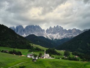 grassy hills with a little hamlet in the center give way to forest and then imposing grey mountains in the Dolomites Italy