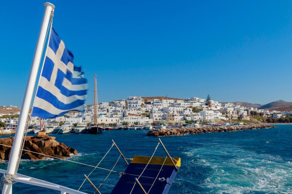 a greek flag waving off the end of a boat as it leaves a village of white-washed houses in Greece