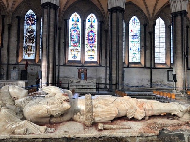 a stone statue lying down with stainedglass windows behind at Salisbury Cathedral