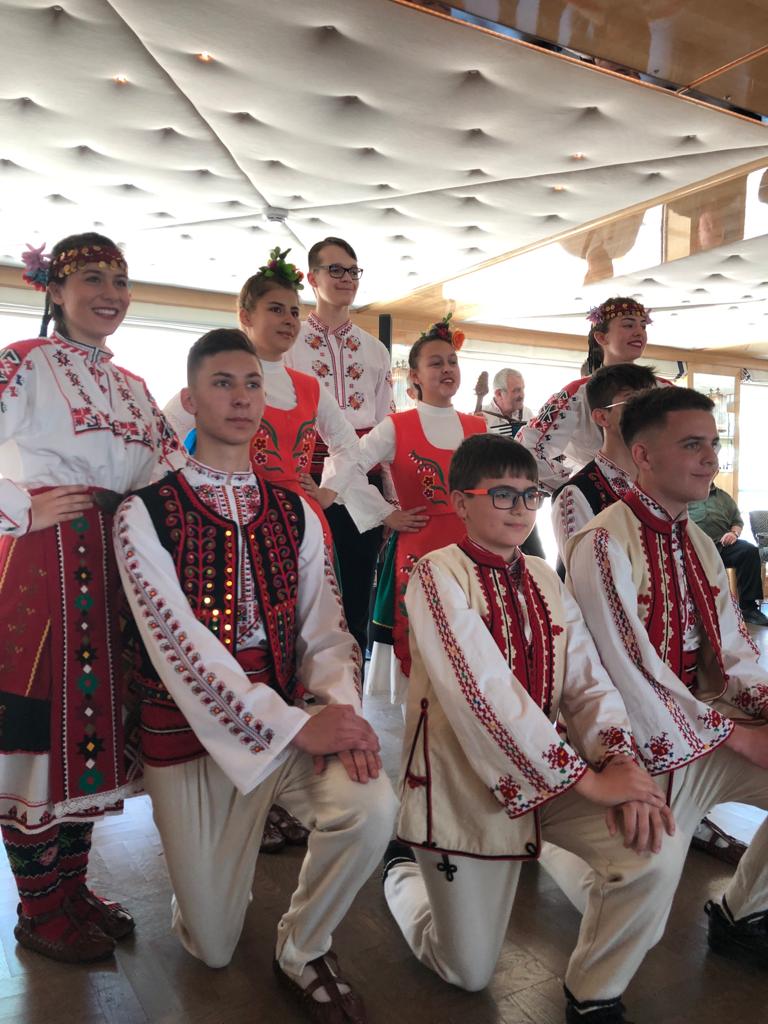 a group of teens and kids in traditional Bulgarian dress kneel for a photo after performing a local folk dance
